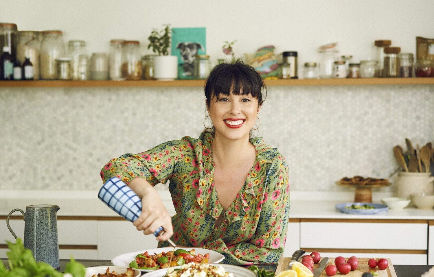 A woman in a kitchen-cooking a mixture of sharing dished-pouring oil-blue and white check olive oil bottle-fresh herbs-chopping fruit and vegetables-open shelf-kitchen units-floral patterned dress