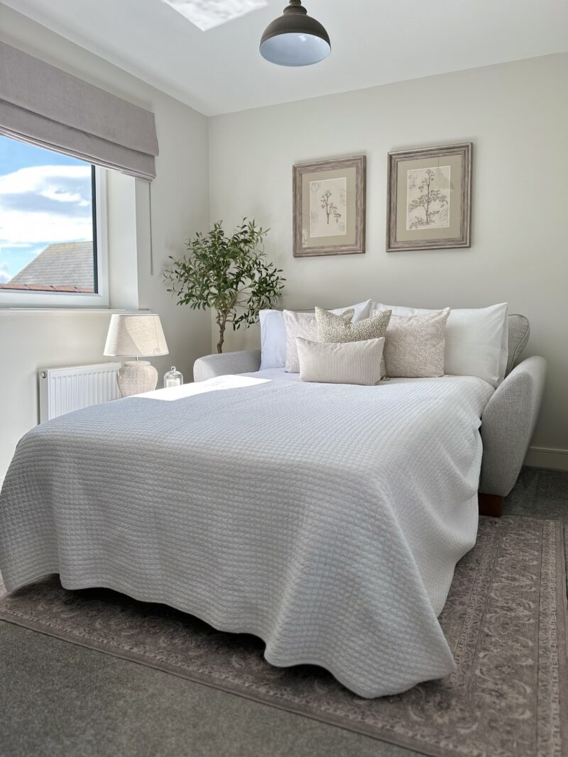 A sofa bed-bedroom furniture-2 seater sofa bed-neutral bedding-vintage style rug-beige carpet-neutral walls-botanical prints-plain blind-cream bedside lamp-brown ceiling light