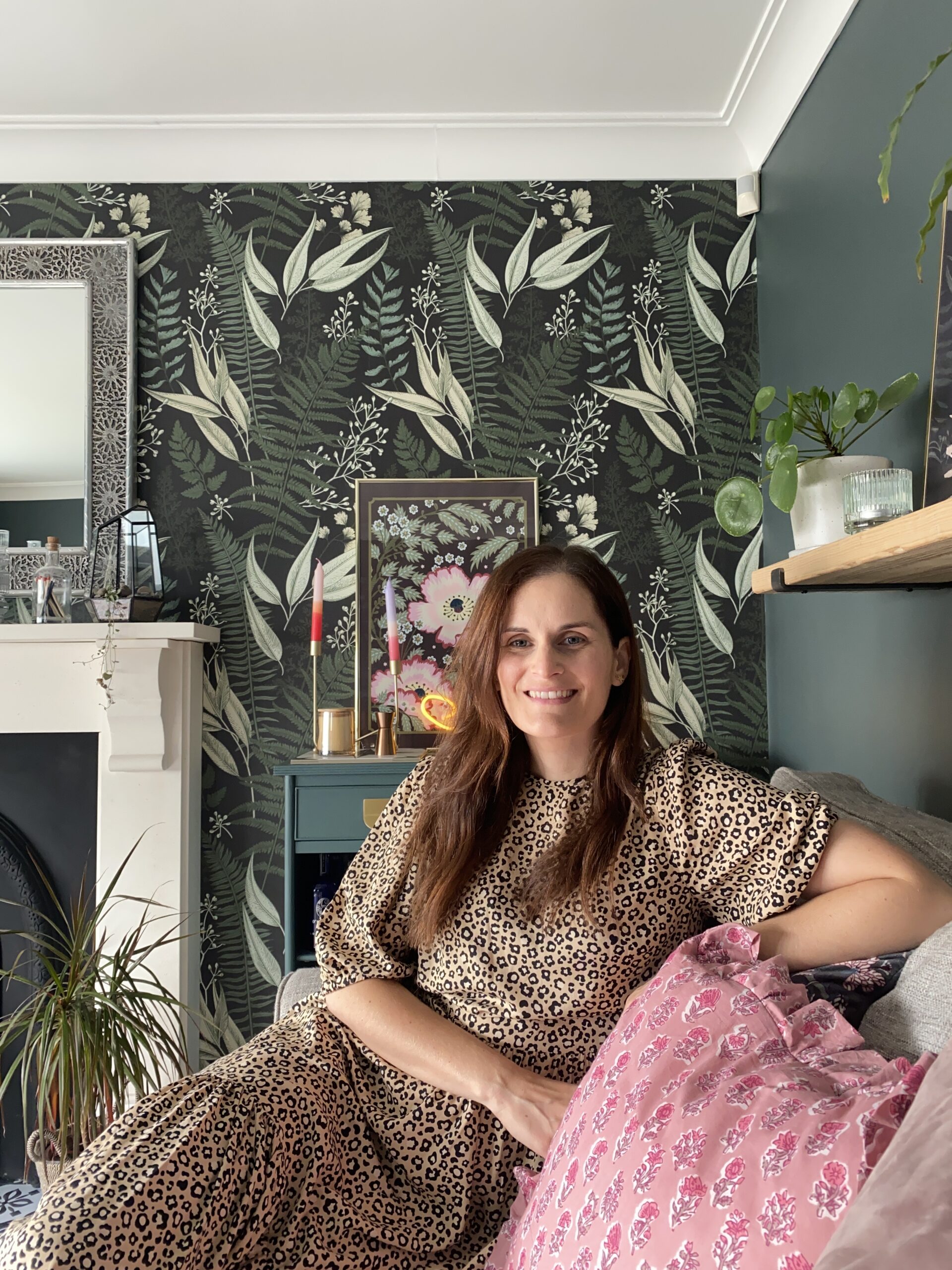 A woman on a sofa-living room furniture-foliage wallpaper-white fireplace-dark green wall-leopard print dress-pink floral cushion-house plants