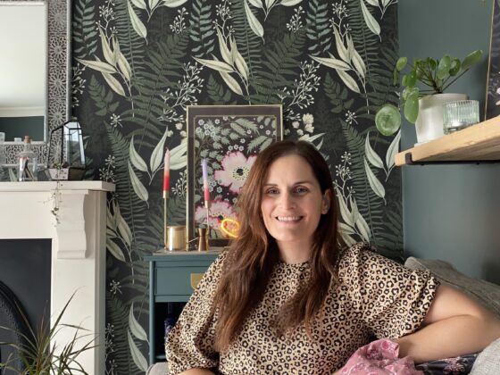 A woman on a sofa-living room furniture-foliage wallpaper-white fireplace-dark green wall-leopard print dress-pink floral cushion-house plants