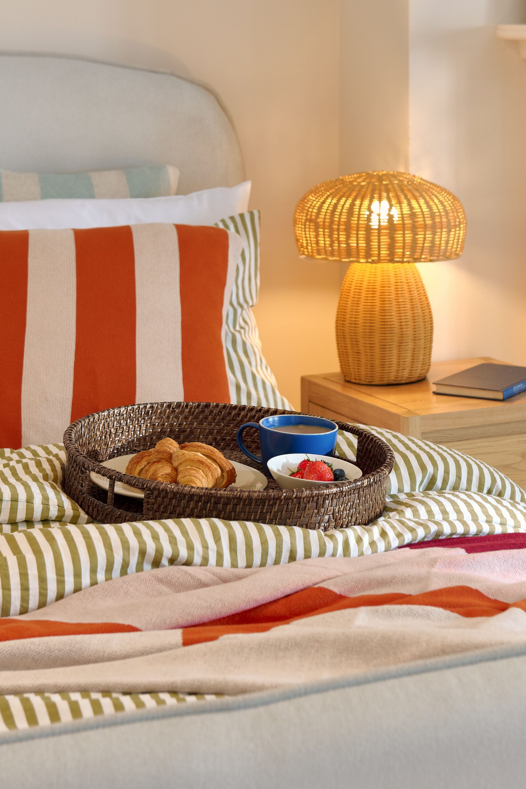 A bed and bedside table-bedroom furniture-green striped bedding-red striped cushion-red and pink throw-brown rattan tray-blue mug of tea-croissants-fresh fruit-neutral upholstered bed-pale wooden bedside table-wicker mushroom lamp