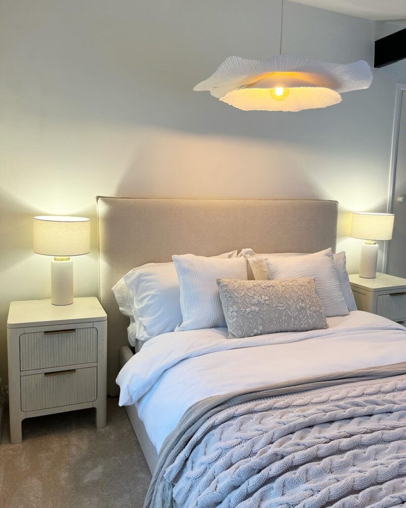 A bed and bedside table-bedroom furniture-white walls-white bedding-neutral knitted throw-Art Deco style bedside table-upholstered bed-bedside table lamps-cloud ceiling light-neutral carpet