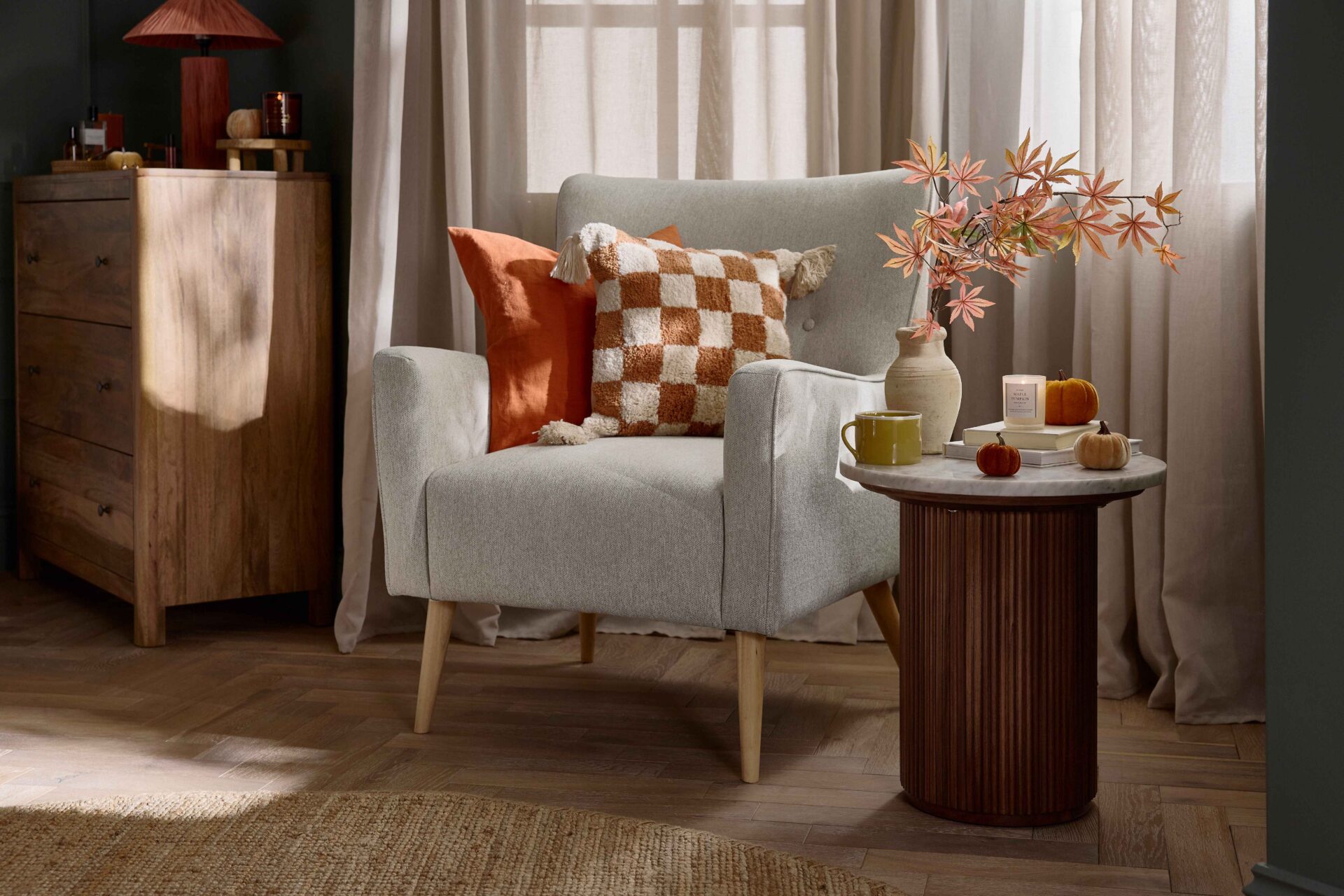 An accent chair and side table-bedroom furniture-marble topped side table-fluted wood-neutral button back armchair-orange check cushion-parquet flooring-sheer draped curtains-round jute rug=mango wood chest of drawers-rich blue walls-velvet pumpkins-autumnal leaves in natural vase
