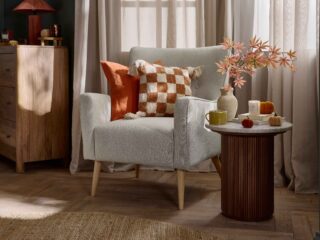 An accent chair and side table-bedroom furniture-marble topped side table-fluted wood-neutral button back armchair-orange check cushion-parquet flooring-sheer draped curtains-round jute rug=mango wood chest of drawers-rich blue walls-velvet pumpkins-autumnal leaves in natural vase