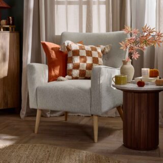 An accent chair and side table-bedroom furniture-marble topped side table-fluted wood-neutral button back armchair-orange check cushion-parquet flooring-sheer draped curtains-round jute rug=mango wood chest of drawers-rich blue walls-velvet pumpkins-autumnal leaves in natural vase