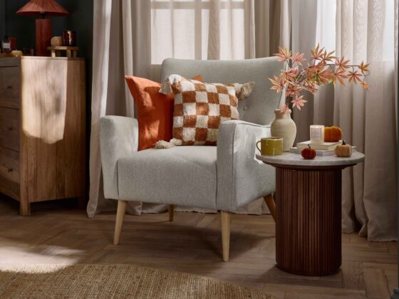 An accent chair and side table-bedroom furniture-marble topped side table-fluted wood-neutral button back armchair-orange check cushion-parquet flooring-sheer draped curtains-round jute rug=mango wood chest of drawers-rich blue walls-velvet pumpkins-autumnal leaves in natural vase