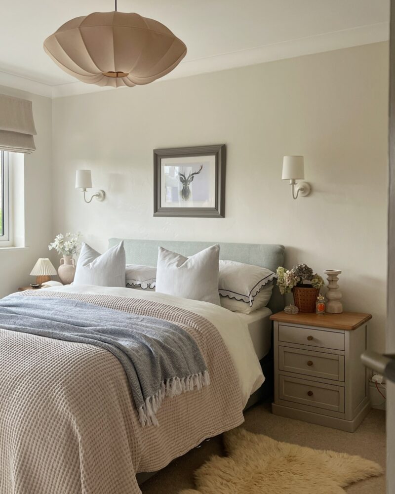 A bed and bedside table-bedroom furniture-neutral walls-upholstered bed-layered bedding-painted wooden bedside table-sheepskin rug-statement ceiling light-delicate wall lights-fresh flowers-natural light