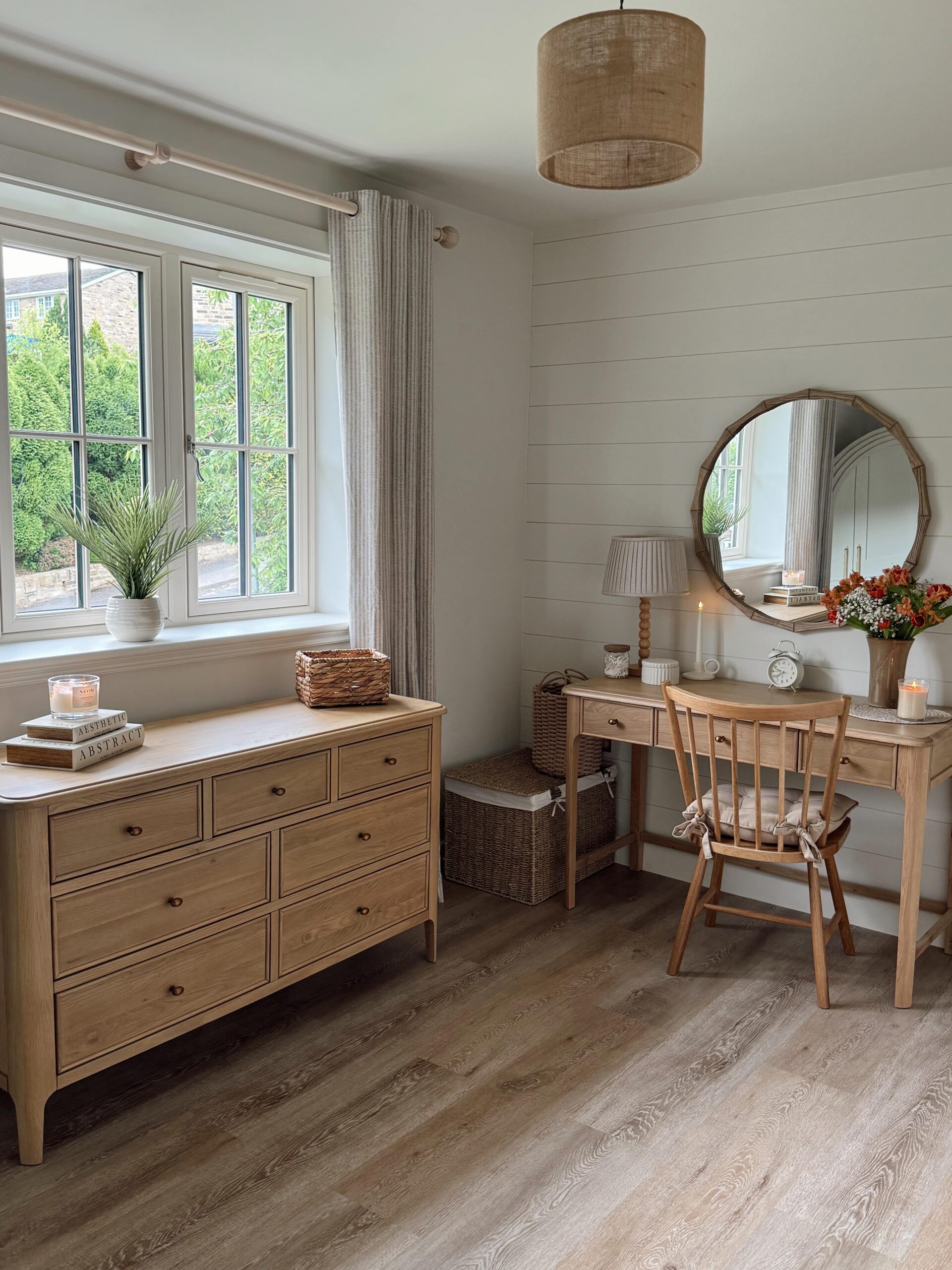 A neutral bedroom corner-natural wood chest of drawers-natural wood dressing table-round bamboo mirror-wooden chair with a cushion-candles-table lamp-clock-vase of fresh flowers-woven storage baskets-large window with light curtains-warm wooden flooring-panelled wall