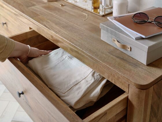 A chest of drawers with hand-bedroom furniture-folded cream trousers-mango wood drawers-storage box-sunglasses-perfume bottles-parquet floor-gold jewellery-stacked books