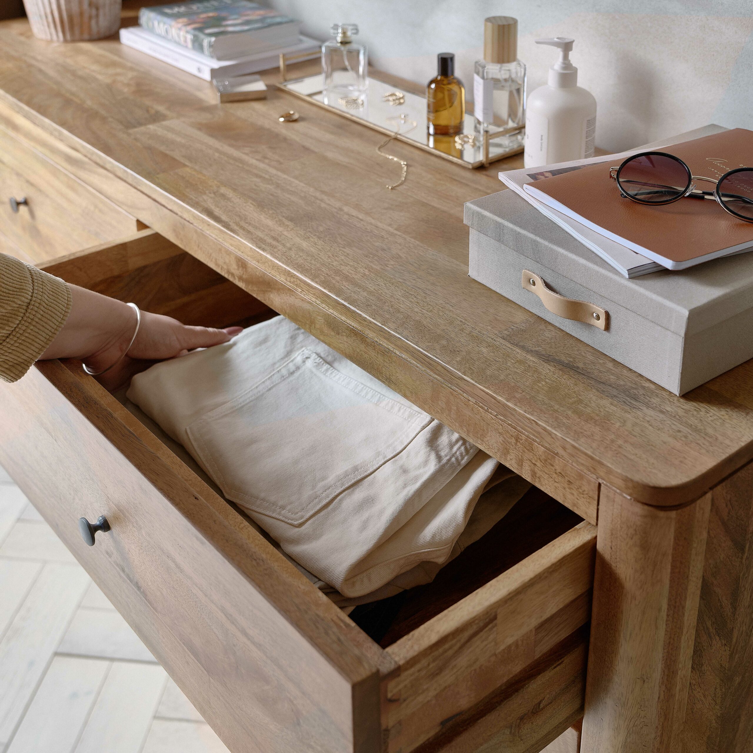 A chest of drawers with hand-bedroom furniture-folded cream trousers-mango wood drawers-storage box-sunglasses-perfume bottles-parquet floor-gold jewellery-stacked books