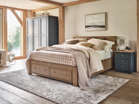 A bed, wardrobe, and bedside table-bedroom furniture-wooden bed with storage drawers-neutral bedding-check bed linen-navy blue cabinetry-wooden floor-textured neutral rug-neutral walls-exposed beams-blue bedside table with drawers-blue triple wardrobe