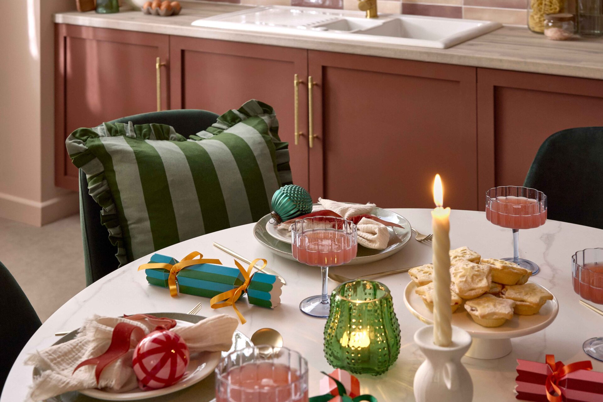 A dining table and chairs-kitchen diner furniture-white ceramic tabletop-green velvet dining chairs-stripey green cushion with frills-christmas tablescape-festive place settings-colourful baubles-striped crackers-twisted candles-green glass tealight-scalloped champagne glasses-burgundy kitchen units-brass door handles