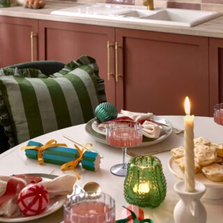 A dining table and chairs-kitchen diner furniture-white ceramic tabletop-green velvet dining chairs-stripey green cushion with frills-christmas tablescape-festive place settings-colourful baubles-striped crackers-twisted candles-green glass tealight-scalloped champagne glasses-burgundy kitchen units-brass door handles