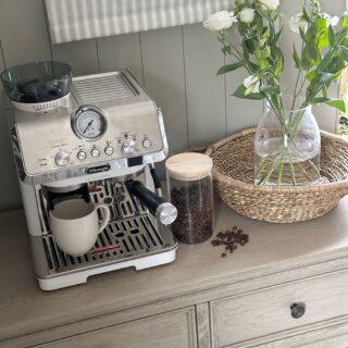 A sideboard-dining room furniture-coffee station-coffee machine-coffee beans in cannister-woven tray-fresh flowers in glass vase-weathered oak sideboard-neutral walls with panelling-striped blind