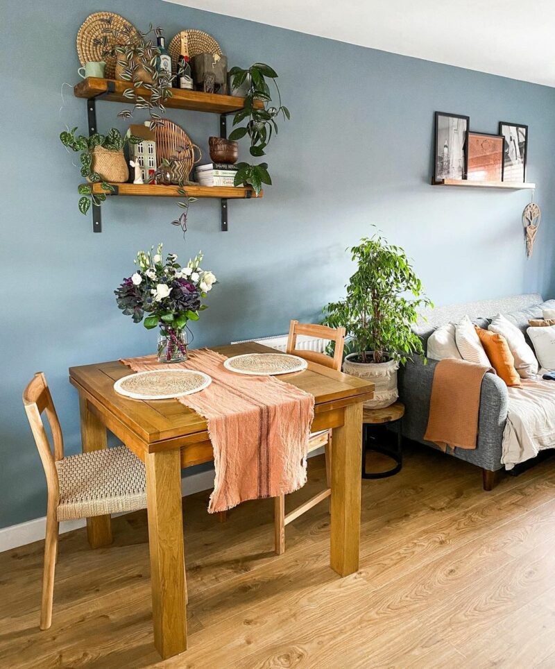 A dining table, chairs, and sofa-dining room furniture-square oak dining table and chairs-wooden wall shelving-grey blue walls-wooden flooring-grey sofa-house plants-vase of flowers-pink table runner