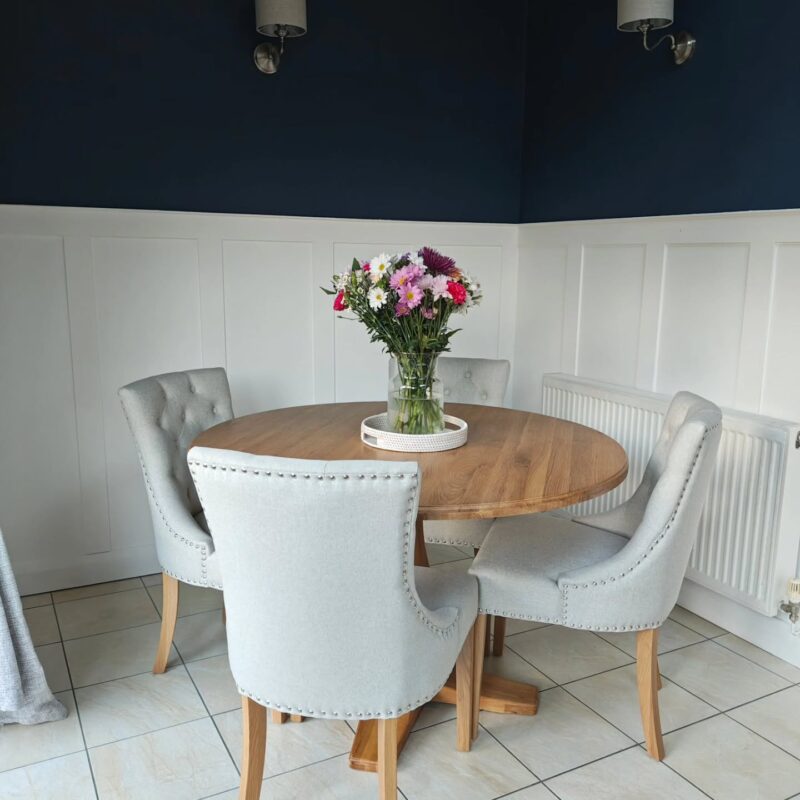 A dining table and chairs-dining room furniture-oak dining table set-round dining table-half wall panelling in white-navy walls-beige tiled floor-glass vase of colourful flowers-grey fabric dining chairs-wall mounted lights