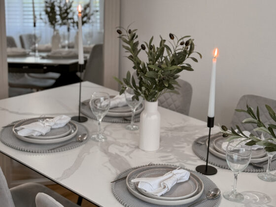A dining table and chairs-dining room furniture-ceramic table-grey upholstered dining chairs-tapered candles-black candle holders-vases with foliage-arched mirror-grey place settings