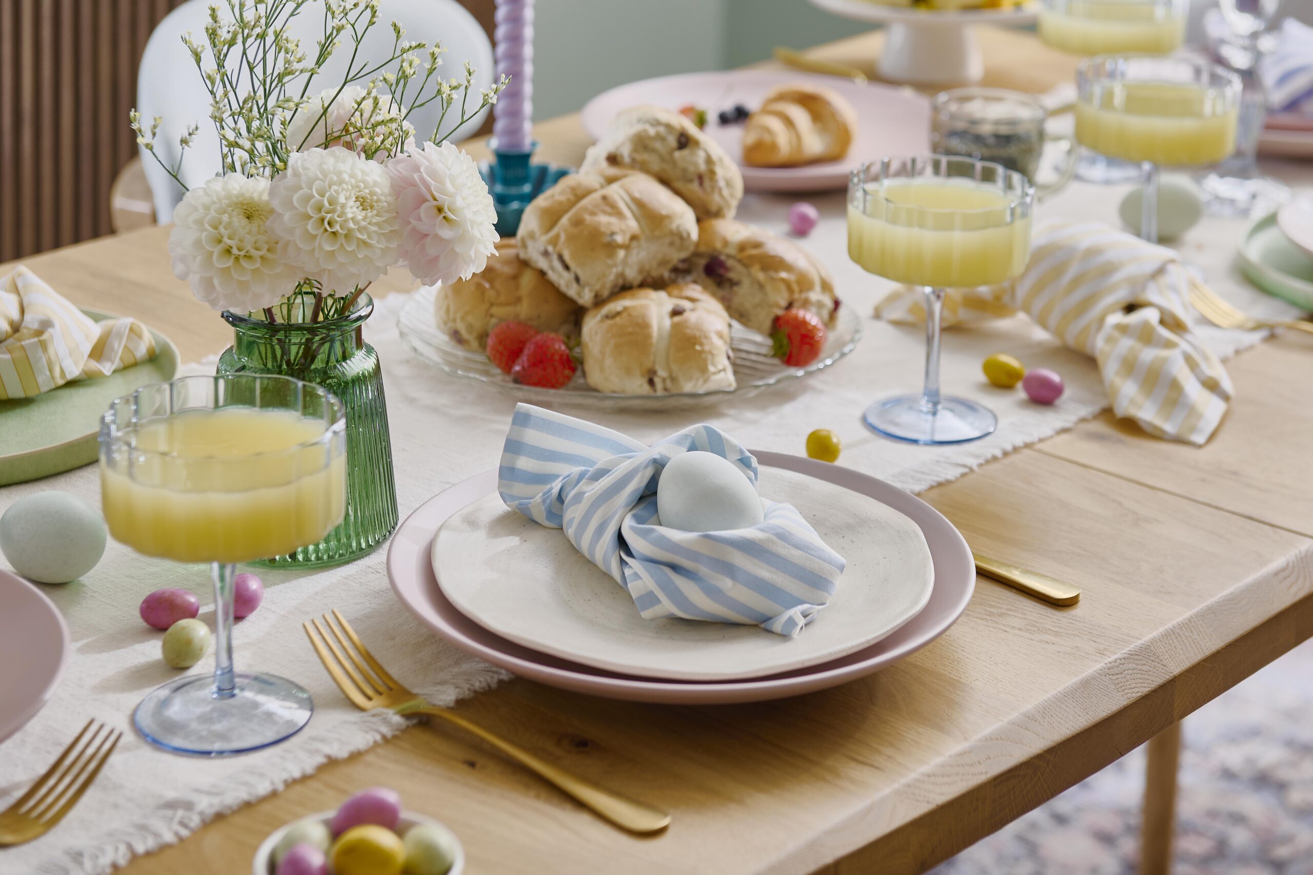 A dining table and chairs-dining room furniture-pale oak wooden table-mid century style upholstered chairs-dark wood sideboard with fluted detail-green blue walls-vintage style rug-colourful tapered candles-Easter tablescape-Easter food-pastel homewares-gold cutlery-coloured glassware-folded striped napkin-duck egg blue eggs