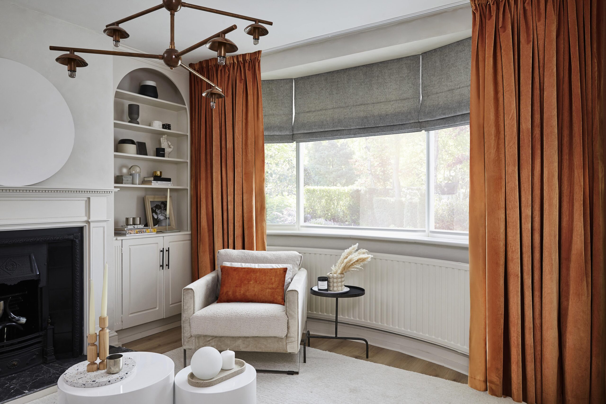 A chair, coffee table and fireplace-living room furniture-orange velvet curtains-grey blinds-statement ceiling light-arched alcove-round plinth coffee tables-off white walls