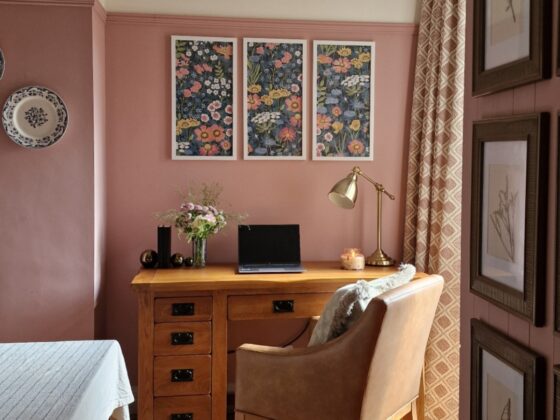 A desk and chair-home office furniture-oak desk with drawers-brown leather look upholstered chair-pink and neutral walls-framed floral wallpaper-botanical artwork-decorative wall plates-patterned curtain-vintage rug-beige tablecloth