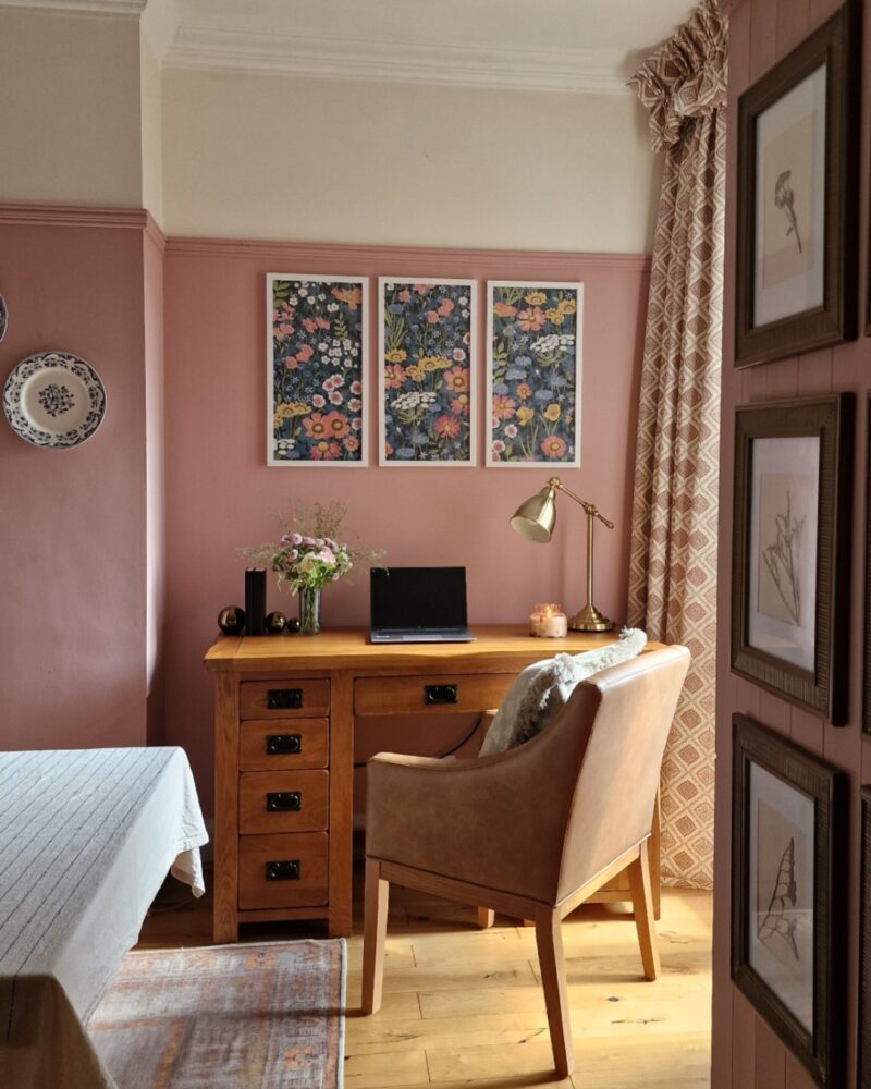 A desk and chair-home office furniture-oak desk with drawers-brown leather look upholstered chair-pink and neutral walls-framed floral wallpaper-botanical artwork-decorative wall plates-patterned curtain-vintage rug-beige tablecloth