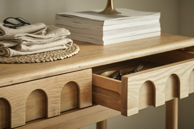 A console table-living room furniture-wooden console table with arch drawer detail-open drawer-linen napkins-black frame glasses-wicker placemat-stacked magazines-neutral walls