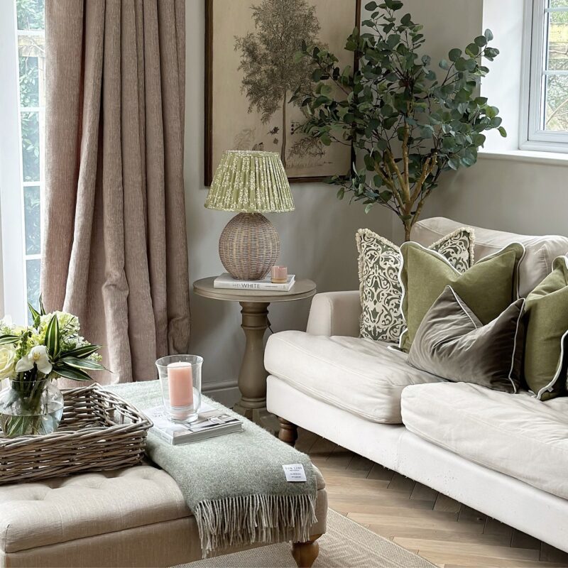 A sofa, side table, and footstool-living room furniture-weathered oak side table-potted plant-foliage artwork-side table wicker lamp-green cushions-woven tray-fresh flowers in glass vase-wool throw-neutral walls-cream sofa