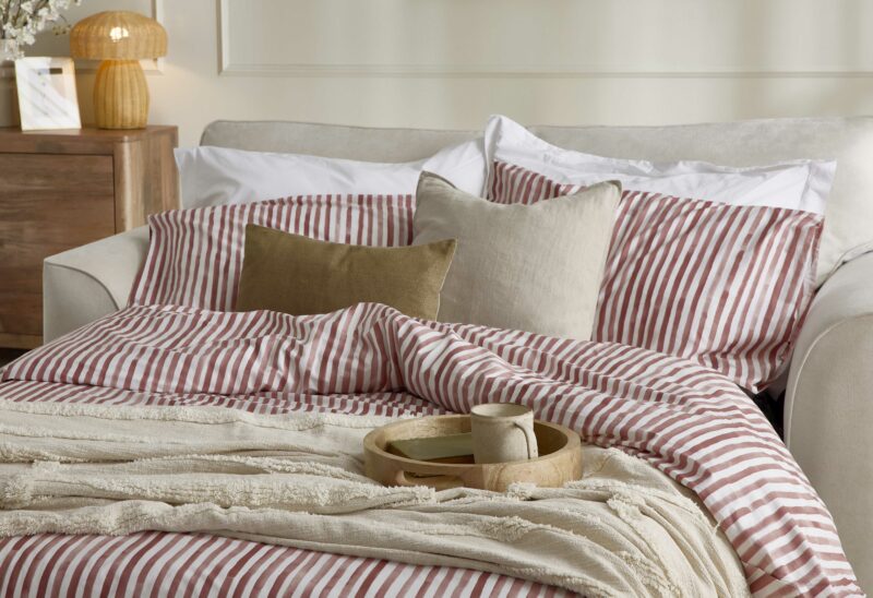 A sofa bed-bedroom furniture-cream 2 seater sofa bed-red striped bedding-neutral throw-beaded walls-wooden tray-rattan mushroom lamp-mango wood sideboard