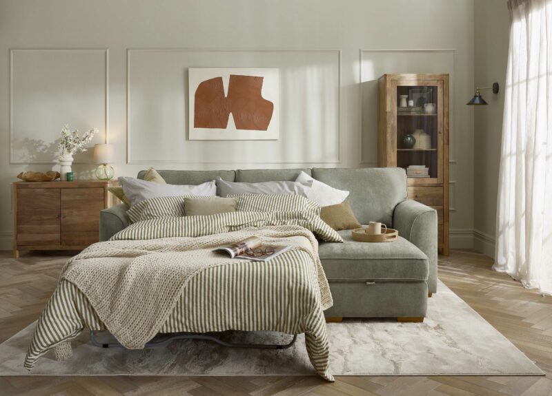 A sofa bed-living room furniture-green corner chaise sofa bed-green striped bedding-neutral knitted throw-beaded walls-wooden tray-glass lamp-mango wood sideboard-display cabinet-abstract wall art-neutral rug-parquet floor-sheer curtain