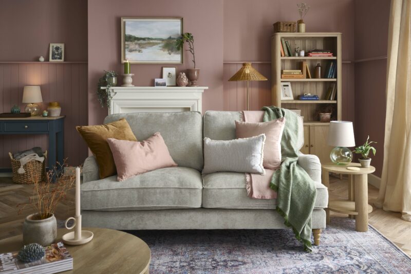A desk, sofa, and bookcase-living room furniture-dark blue painted desk-dusky pink walls-parquet wooden floor-vintage pattern rug-woven storage basket-grey sofa-tan velvet cushion-pink linen cushion-fireplace-potted plants-landscape wall art-pale oak coffee table-round oak side table