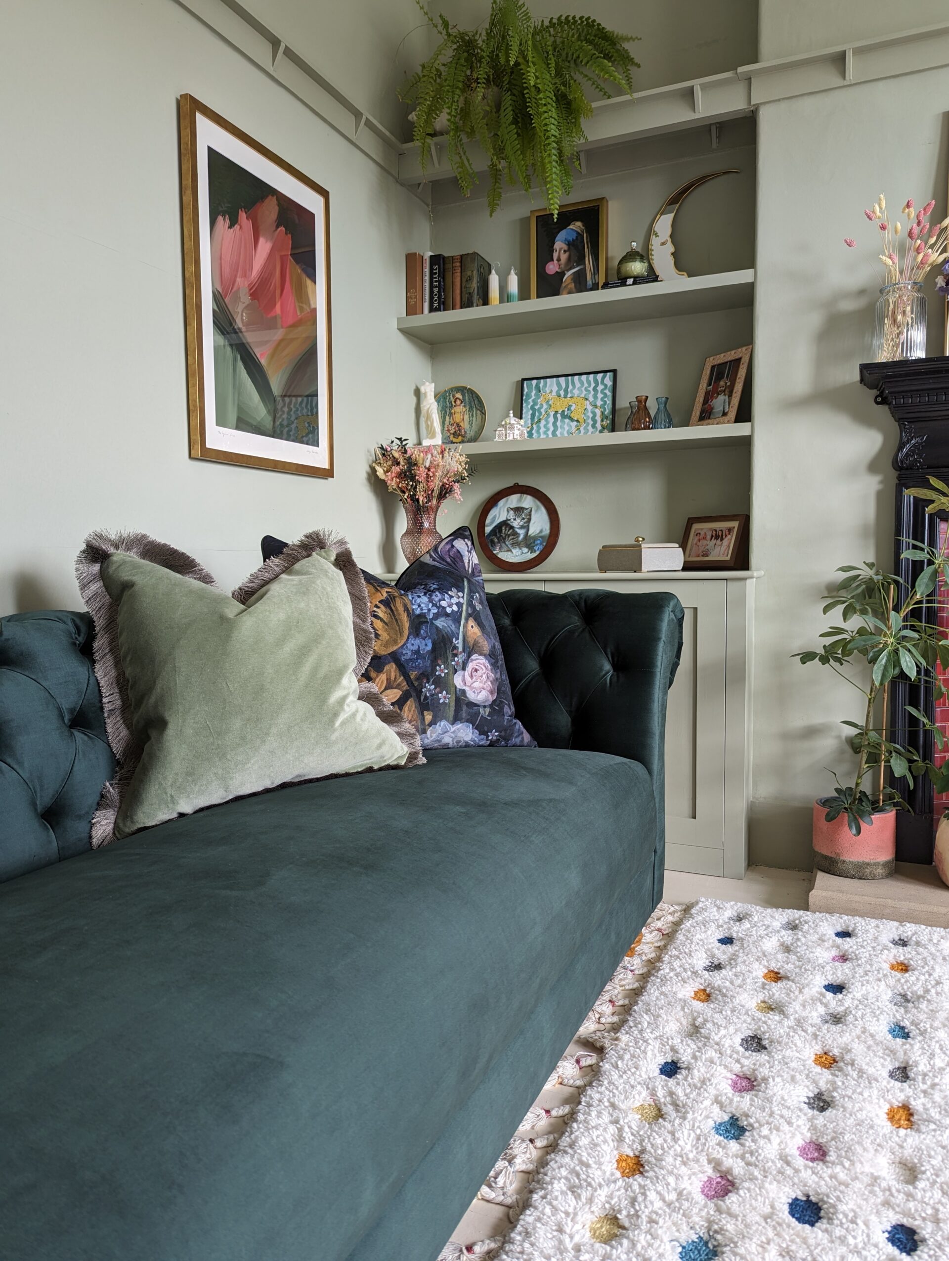 A sofa-living room furniture-plush velvet green sofa-velvet cushion-patterned cushion-soft green walls-polka dot rug-built in shelving-wall art-fireplace-house plants