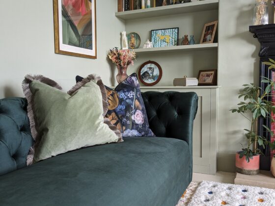 A sofa-living room furniture-plush velvet green sofa-velvet cushion-patterned cushion-soft green walls-polka dot rug-built in shelving-wall art-fireplace-house plants
