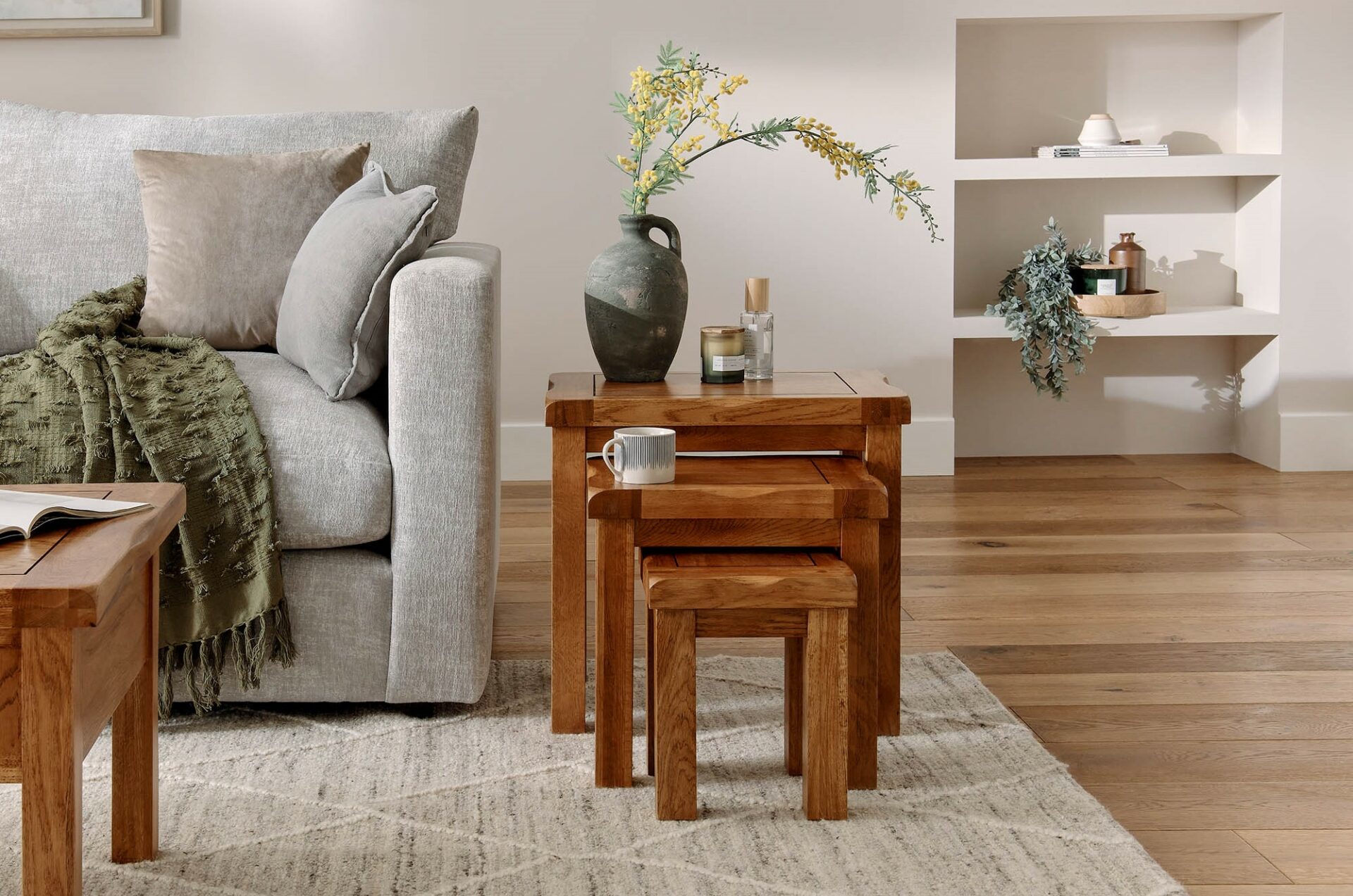 A nest of tables and sofa-living room-wooden nest of tables-a soft grey sofa-neutral cushions-a green throw-a ceramic vase with yellow flowers-a candle-a mug-built in shelves-plants-books-wooden flooring-patterned rug