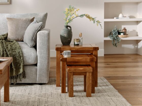 A nest of tables and sofa-living room-wooden nest of tables-a soft grey sofa-neutral cushions-a green throw-a ceramic vase with yellow flowers-a candle-a mug-built in shelves-plants-books-wooden flooring-patterned rug