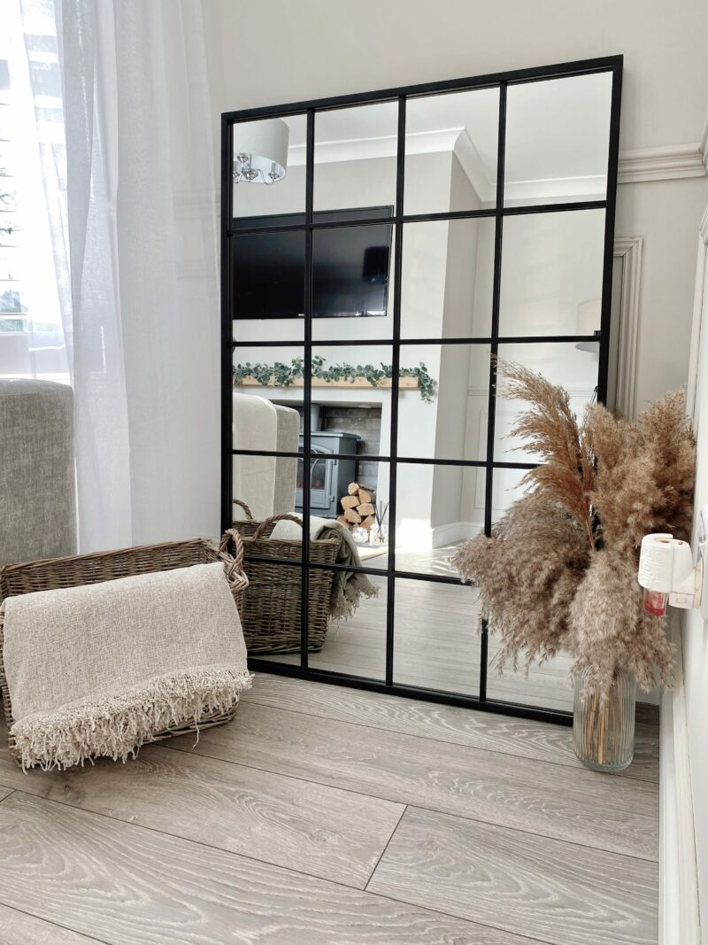 A mirror-living room furniture-black windowpane floor mirror-dried grass in glass vase-wicker storage basket-cream throw-neutral walls-log burner-white washed wooden floorboards