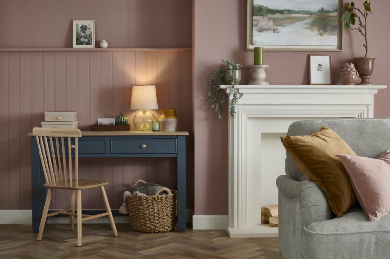 A desk, chair, and sofa-living room furniture-dark blue painted desk-wooden spindle-back chair-dusky pink walls-parquet wooden floor-woven storage basket-grey sofa-tan velvet cushion-pink linen cushion-fireplace-potted plants-landscape wall art