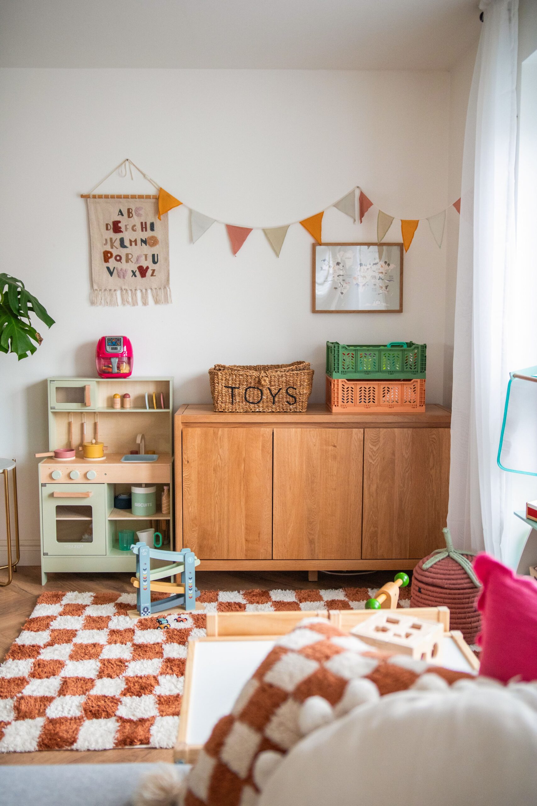 A sideboard-playroom furniture-wooden sideboard-storage cabinet-patterned checkerboard rug-play kitchen-storage crates-wicker baskets-colourful bunting