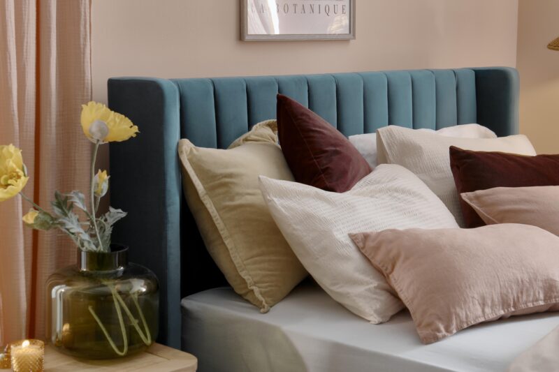A bed and bedside table-bedroom furniture-teal upholstered bed-layered cushions-plain mattress-pink walls-green vase with flowers-candle light-cream and wood bedside table-floral wall art