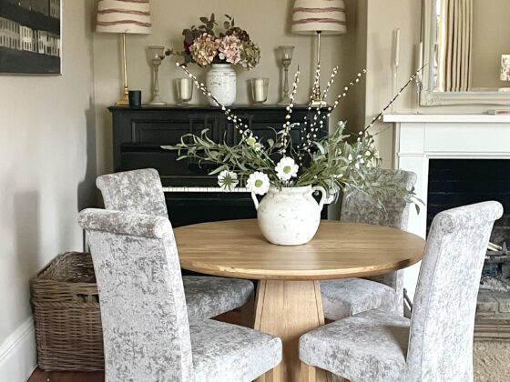 Round oak table with grey upholstered chairs in an elegant living room with a piano in the background.